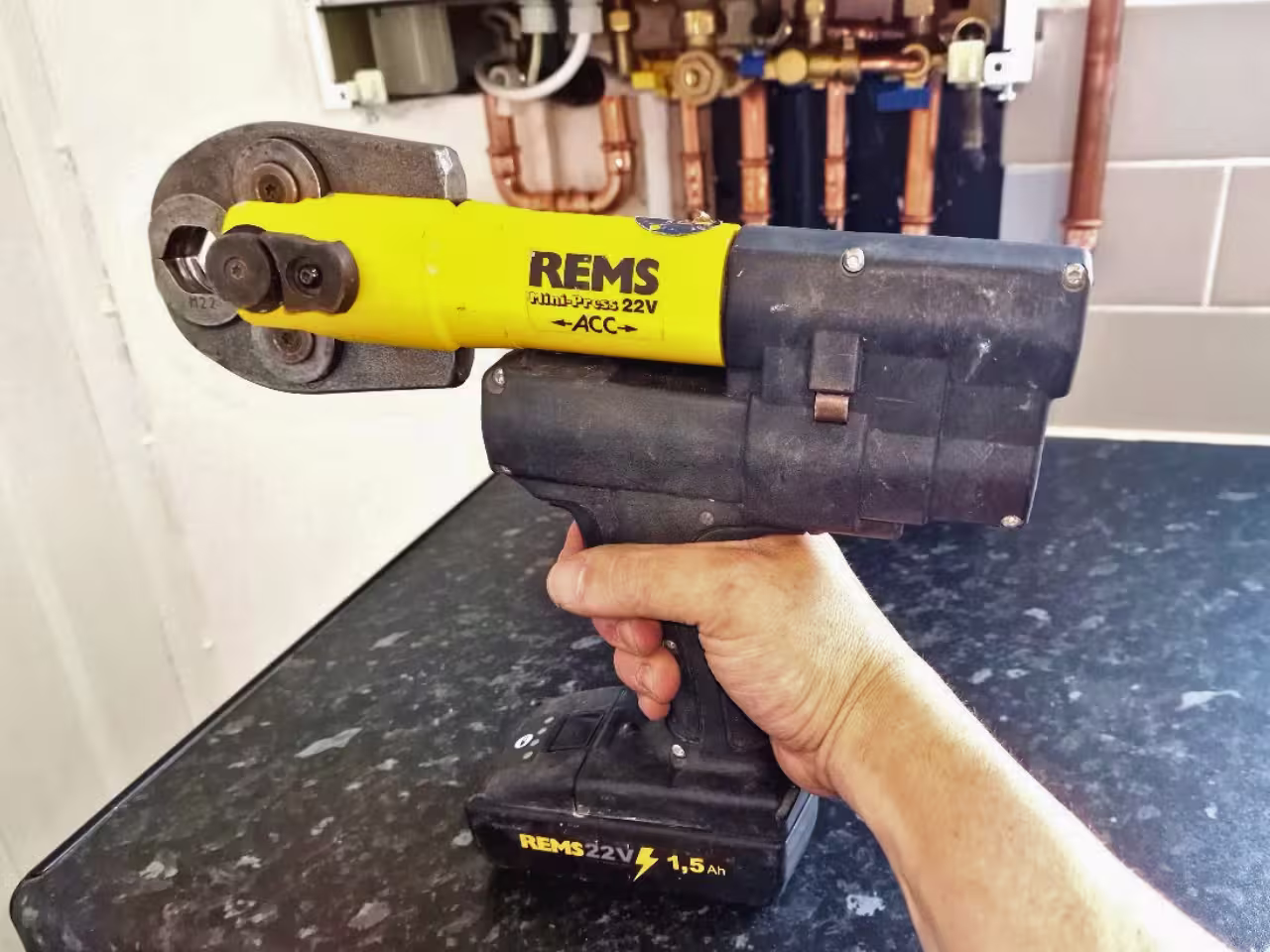 REMS Mini-Press 22V ACC held in front of a boiler with copper pipework